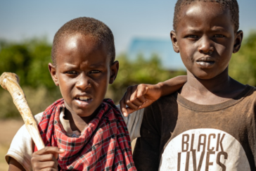 Zwei Jungs im kenianischen Dorf. Sie sehen aus, als hätte man sie gerade beim Spielen etwas gestört, aber sie geben gerne ein gutes Motiv ab. Der linke Junge hat einen Stock über der Schulter liegen.