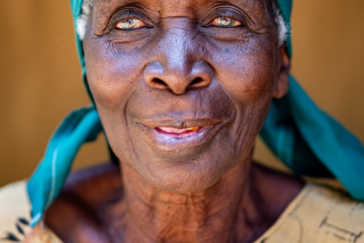 Ein Porträt einer alten Kenianerin. Sie trägt ein strahlend türkises Tuch auf dem Kopf und ein beiges Kleid mit zartem Blütenmuster. Ihre Augen sind schon etwas trüb, aber sehr ausdrucksstark.
