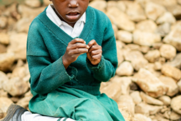 Ein Kind in Schuluniform in Kenia. Es sitzt auf einem Stein und schaut in die Kamera. Sein Blick ist interessiert und skeptisch zugleich.