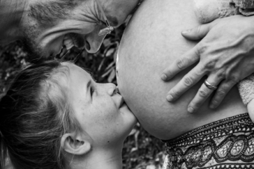 Eine dreiköpfige Familie bekommt Nachwuchs: In dieser Schwarz-Weiß-Aufnahme küsst ein kleines Mädchen den Babybauch der Mutter und der Vater hat die Stirn auf dem Bauch. Sein lächelndes Gesicht schwebt ganz knapp über dem Kopf seiner erstgeborenen Tochter.