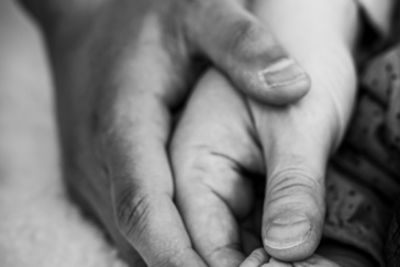 Hände von Eltern und Baby, der Vater hält die Hand der Mutter, die Mutter die Hand des Babys. Die Aufnahme ist in Schwarz-Weiß.