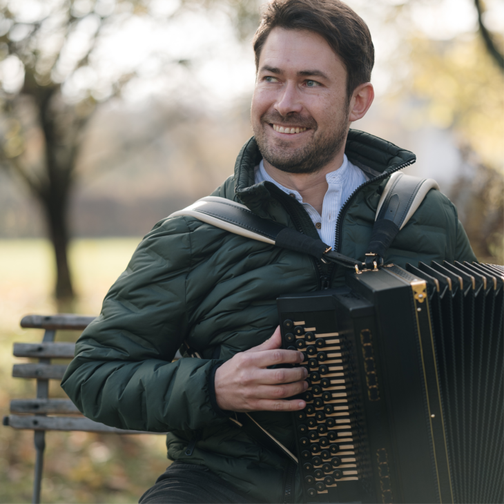 Johannes Sift sitzt mit seiner Harmonika aus dunklem Holz in der Herbstsonne draußen auf einer Bank.